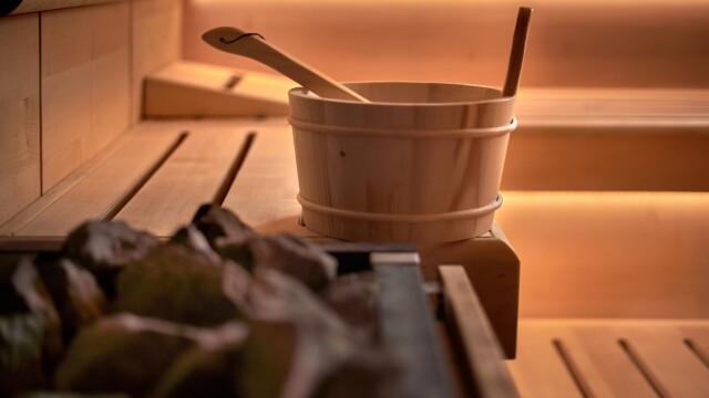 A wooden sauna room with a bench, a bucket, a ladle, and hot stones, softly lit by warm ambient lighting.