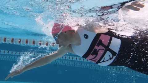 Underwater view of a swimmer in a pink swim cap and patterned swimsuit, reaching forward with one arm while swimming freestyle in a clear blue pool near a lane divider.