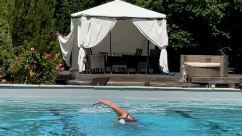 A person swims freestyle in a pool in front of a white outdoor canopy with curtains, chairs, and a table. Lush greenery and trees surround the scene, giving it a relaxing, summery atmosphere.
