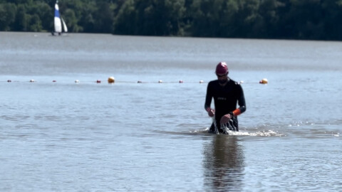 A person in a wetsuit and swim cap walks through shallow lake water, splashing slightly, with a line of buoys behind them and trees and a sailboat visible in the background.