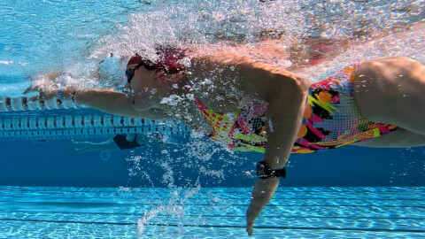 A swimmer in a colorful swimsuit and swim cap does a front crawl stroke underwater in a pool, with bubbles and water movement surrounding her. The pool’s blue tiles and lane divider are visible.