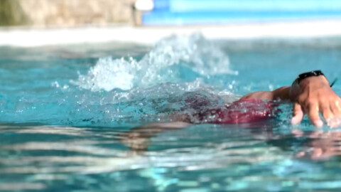 A person wearing a red swimsuit is swimming in a pool, creating splashes with their arm as they move through the water. The focus is on their arm and the water surface.