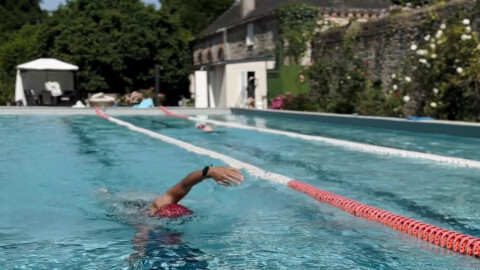 A swimmer wearing a red swim cap and goggles swims freestyle in an outdoor pool, separated by lane dividers, with buildings and greenery in the background on a sunny day.