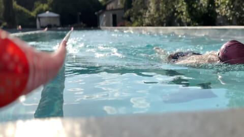 A person swimming in an outdoor pool is seen from water level; another hand in the foreground holds a floating object, pointing toward the swimmer. Trees and buildings are visible in the background.