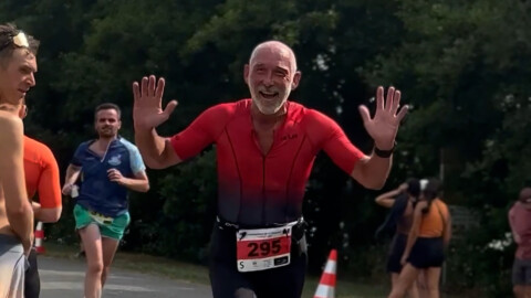 An older man wearing a red shirt with race number 295 smiles joyfully and raises both hands while running in a race. Other runners and a traffic cone are visible in the background.
