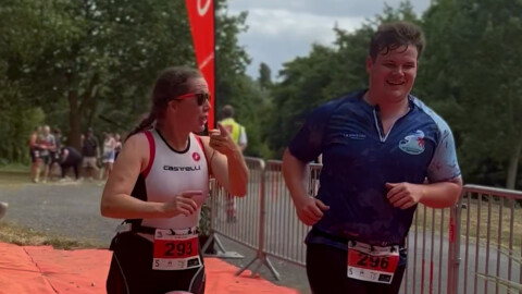 Two athletes wearing race bibs, one in a white Castelli tri-suit and the other in a blue top, run side by side past a finish line, smiling and sweating, with other participants in the background.