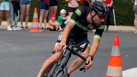 A cyclist wearing a helmet and sunglasses makes a sharp turn on a road during a race, with orange traffic cones and spectators watching from the side.