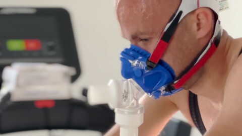 A person sweats while wearing a blue and white respiratory mask connected to medical equipment, possibly during a fitness or breathing test. A treadmill is blurred in the background.