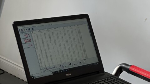 A Dell laptop displaying a data spreadsheet is placed on the control panel of a treadmill, with sunlight illuminating part of the scene. The treadmill’s armrests and a portion of the console are also visible.