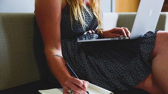 A woman in a patterned dress sits on a couch, holding a laptop on her lap and writing in a notebook with a pen. Her long hair is loose, and her focus is on her notebook.
