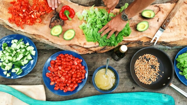Two people prepare fresh vegetables, including lettuce, avocado, tomatoes, cucumber, and red pepper, on a rustic wooden board, surrounded by bowls of ingredients and a pan with corn on a wooden table outdoors.