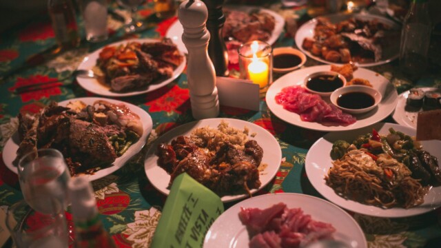 A table covered with various plates of food, including meats, noodles, rice, and sushi, lit by a candle and decorated with festive colors. A salt and pepper grinder are in the center of the table.