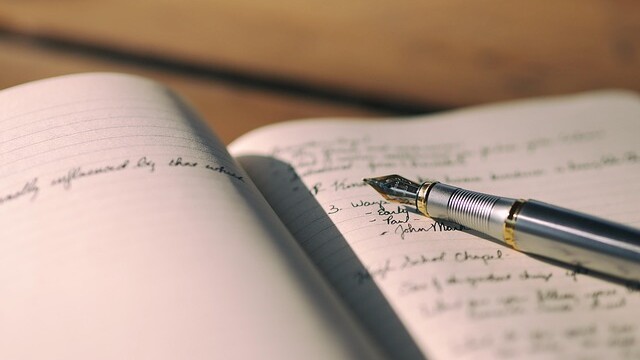 A close-up of an open notebook with handwritten notes and a silver fountain pen resting on the pages. The background is softly blurred, suggesting a calm and focused writing environment.