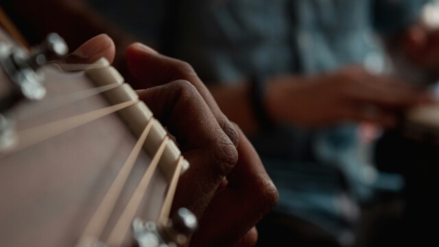 A close-up of a person’s hand playing an acoustic guitar, with another person blurred in the background, possibly playing a percussion instrument. The focus is on the guitar strings and fingers.