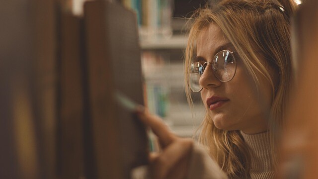 A young woman with glasses and long blonde hair looks closely at books on a shelf in a library, reaching out to select one.