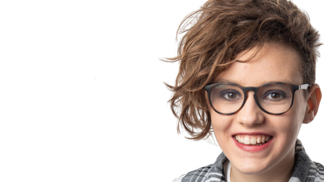 Person with short, wavy brown hair and black-rimmed glasses smiling at the camera, wearing a plaid shirt, against a white background—ready for a creative photography workshop or holiday adventure.