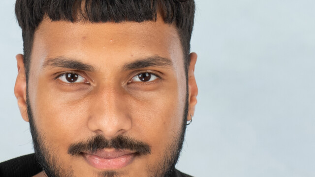 A close-up of a young man with short dark hair, a trimmed beard, and mustache, wearing a small hoop earring and black collar, gazing directly at the camera against a light background—perfect inspiration for creative photography.
