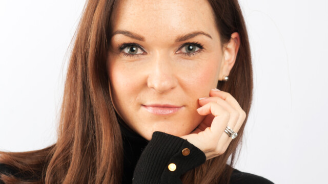 A woman with long brown hair and green eyes, wearing a black top with gold buttons, rests her chin on her hand and looks directly at the camera—capturing a moment of creative photography against a plain white background.