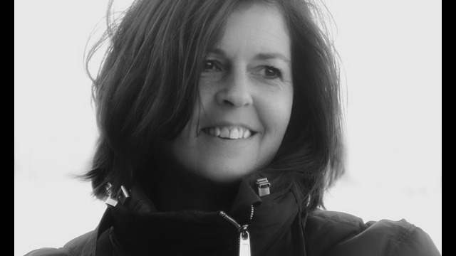 A woman with shoulder-length dark hair smiles while wearing a zipped-up, puffy jacket. The bright, blurred background creates a soft-focus effect, capturing the warmth often seen in creative photography.